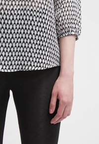 Transparent svartvit topp med ett upprepande skalsmönster, med samlade ärmar och ihop med svarta leggings.