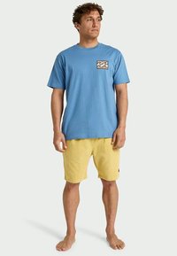 Hombre de pie descalzo, vistiendo una camiseta azul con un logo de ola en el pecho y pantalones cortos amarillos hasta la rodilla, contra un fondo sencillo.