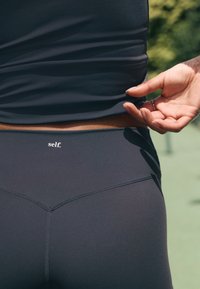 Črna športna majica in pajkice iz gladkega elastičnega materiala. Pajkice imajo šivano šiv in majhen logotip "self." na pasu.