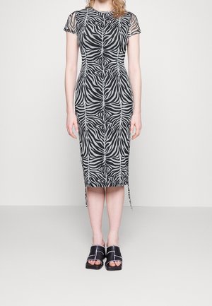 Kobieta w dopasowanej sukience do kolan z czarno-szarym wzorem zebry i przezroczystymi krótkimi rękawami, zestawiona z czarnymi sandałami na otwartym palcu.