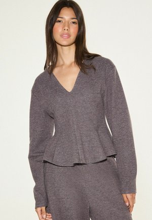 Vrouw draagt een bijpassende zachte grijze gebreide V-hals top met lange mouwen en broek, staand tegen een effen lichte achtergrond.