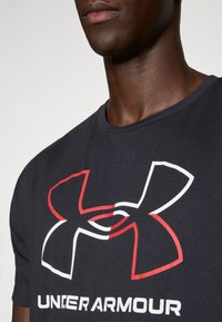 Svart bomullströja med en stor Under Armour-logotyp i vitt och rött. Rund halsringning med en slät textur som syns del av en hals och hak.