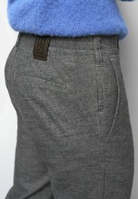 Nahaufnahme von grauen, strukturierten Hosen mit Seiten- und Gesäßtaschen, einem schwarzen Lederlabel, getragen mit einem blauen Strickpullover.