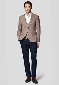 Brązowa marynarka w kratę na białej koszuli, zestawiona z granatowymi spodniami i brązowymi skórzanymi butami. Dopasowany krój, pojedyncze zapięcie na guzik i kołnierz w kształcie ostrego.