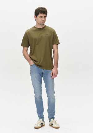 Olive green short-sleeve t-shirt with a round neckline and black logo, paired with light blue slim-fit jeans and off-white sneakers with black stripes.