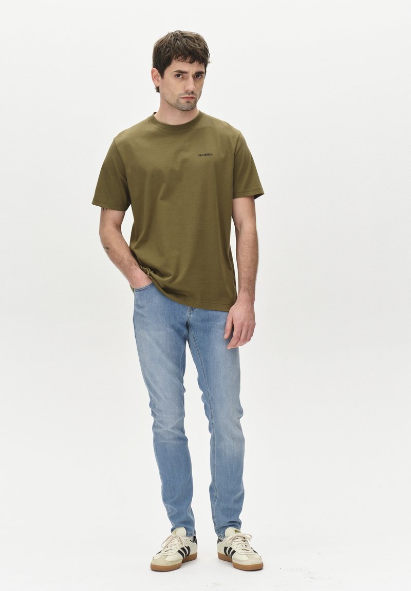 Olivovo zelené tričko s krátkym rukávom a okrúhlym výstrihom, doplnené čiernym logom, v kombinácii so svetlými modrými slim-fit džínsami a krémovými teniskami s čiernymi pruhmi.