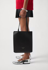 Svart skinn tote bag med en jevn tekstur, lange håndtak og en gull logo. Modellen har på seg metallisk sølvfargede ballerinas og en ribbestrikket rød genser.
