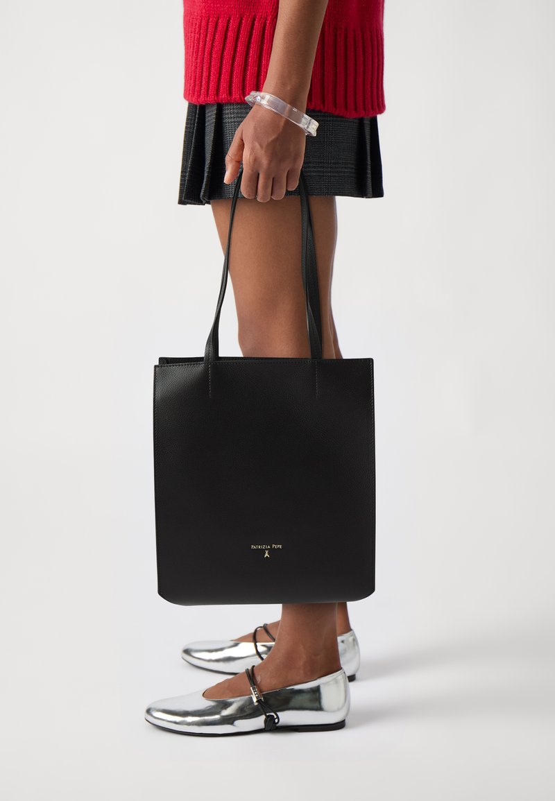 Svart skinn tote bag med en jevn tekstur, lange håndtak og en gull logo. Modellen har på seg metallisk sølvfargede ballerinas og en ribbestrikket rød genser.