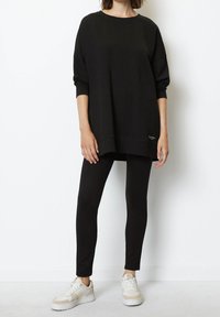 Persona vestida con un top negro de manga larga y oversized, leggings negros ajustados y zapatillas blancas, de pie contra una pared blanca y lisa.