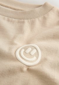 Pull en maille beige avec col rond côtelé et emblème blanc en relief d'un visage souriant sur la poitrine.