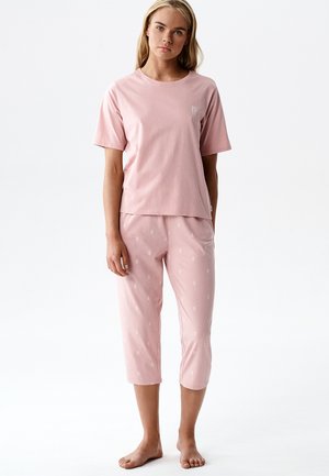 Kobieta stoi boso, ubrana w jasnoróżową koszulkę z krótkim rękawem i dopasowane skrócone spodnie z subtelnymi białymi wzorami.