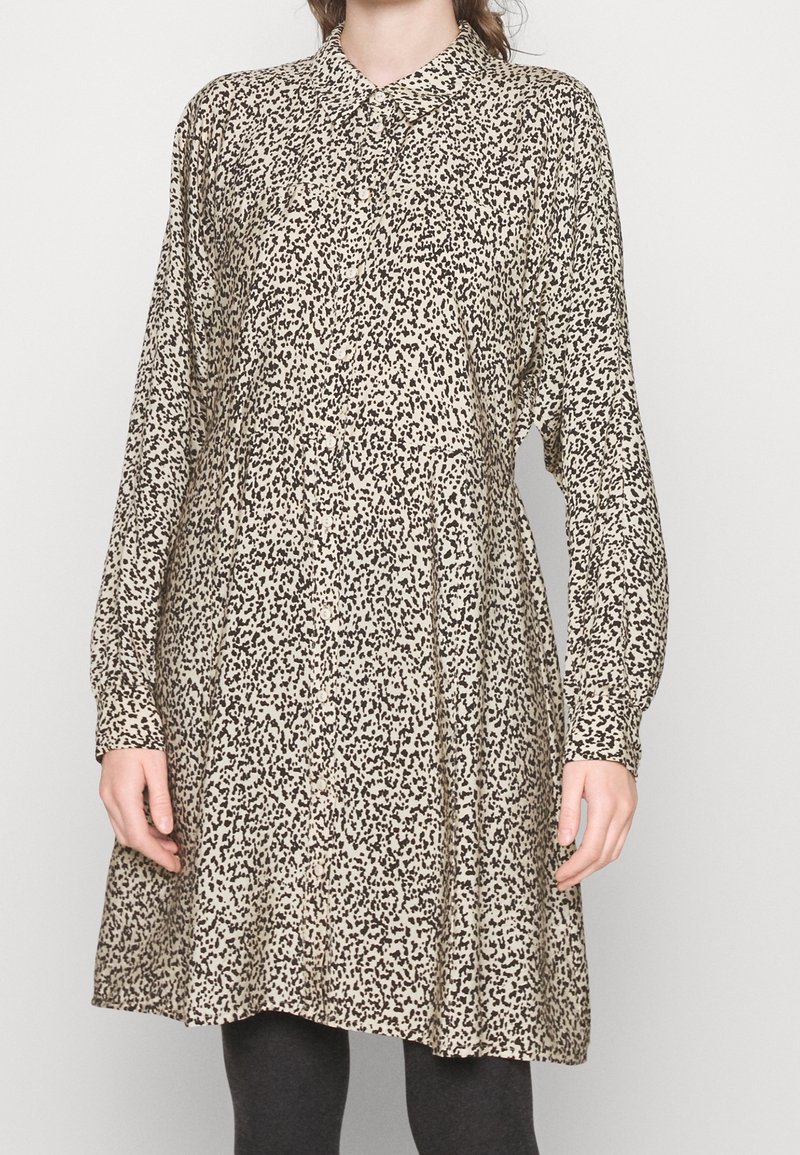 Femme portant une robe beige ample à manches longues avec un motif de points noirs irréguliers et une patte de boutonnage à l'avant, associée à des leggings foncés.