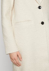 Abrigo de color crema con tejido texturizado y peludo. Presenta botones negros y un bolsillo lateral. Se ve una mano apoyada en la manga del abrigo.