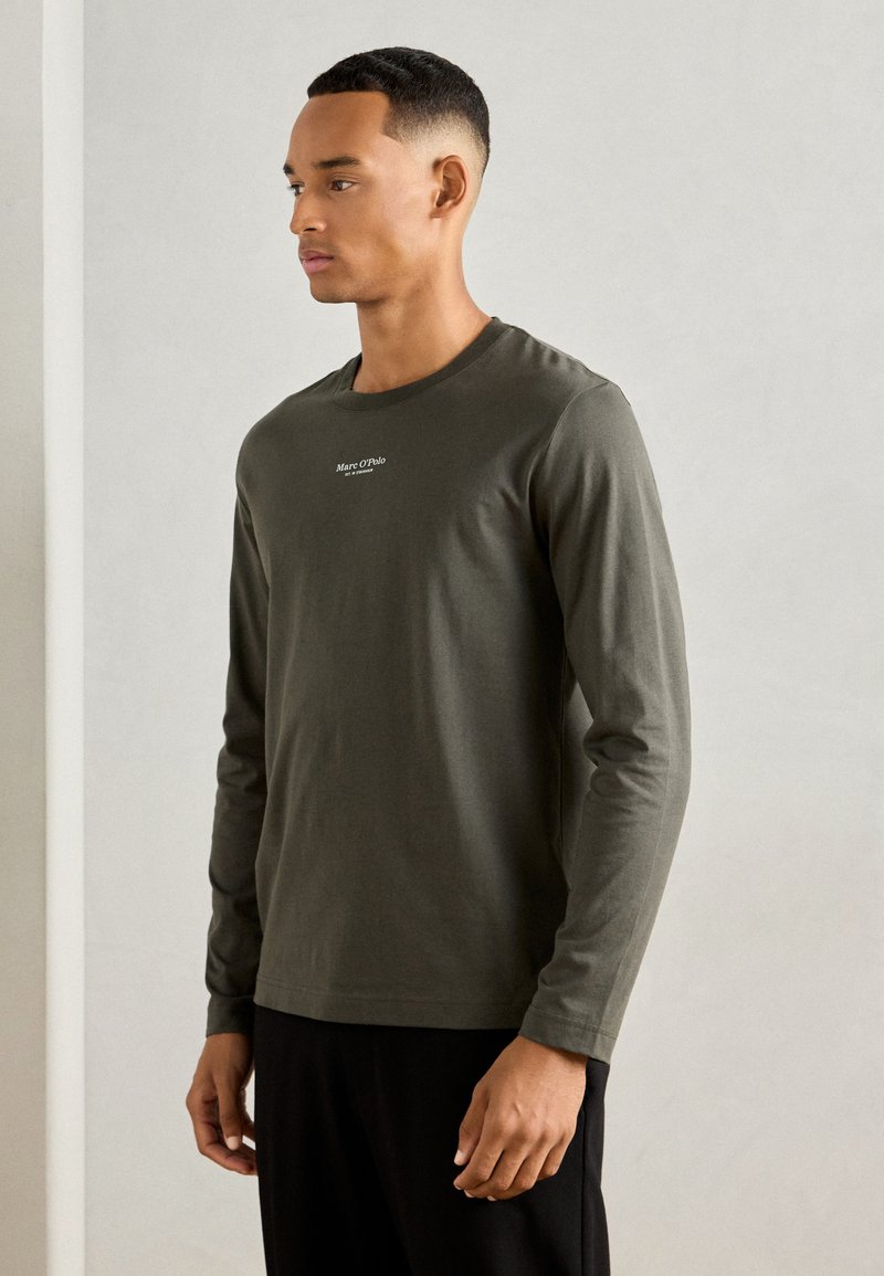 Camicia a maniche lunghe di colore verde oliva in tessuto morbido, con collo a giro e piccolo logo bianco sul petto, abbinata a pantaloni neri.