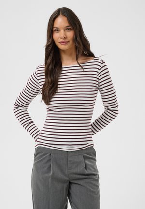 Femme aux longs cheveux foncés portant un haut à manches longues blanc rayé de rouge foncé et un pantalon gris, debout devant un fond uni.