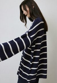 Pull en bleu marine avec des rayures blanches horizontales, texture côtelée, coupe décontractée et manches larges, mettant en avant un design relax.