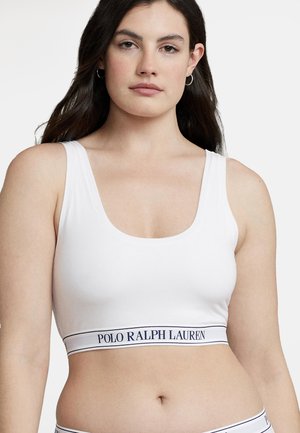 Kobieta o długich ciemnych włosach, ubrana w biały sportowy stanik z napisem "Polo Ralph Lauren" na elastycznej taśmie, zwrócona przodem, o neutralnym wyrazie twarzy.