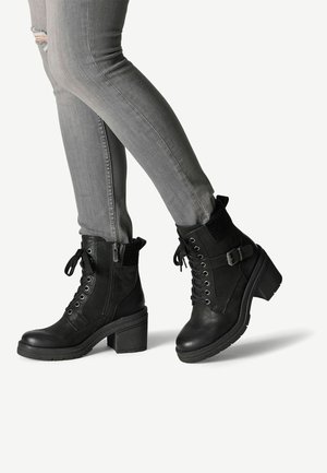 Bottines noires en cuir texturé avec un laçage sur le devant, une fermeture éclair sur le côté et un talon carré ; associées à un jean gris ajusté avec des genoux déchirés.