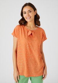 Oranje stoffen top met korte mouwen en een strik bij de halslijn, met gouden rechthoekige patronen erdoorheen. Lichtgewicht en relaxte pasvorm.