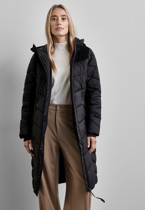 Zwarte pufferjas met een lange snit, voorzien van een gewatteerde textuur, capuchon en zijvakken. Draag het over een lichtgrijze trui en beige broek.