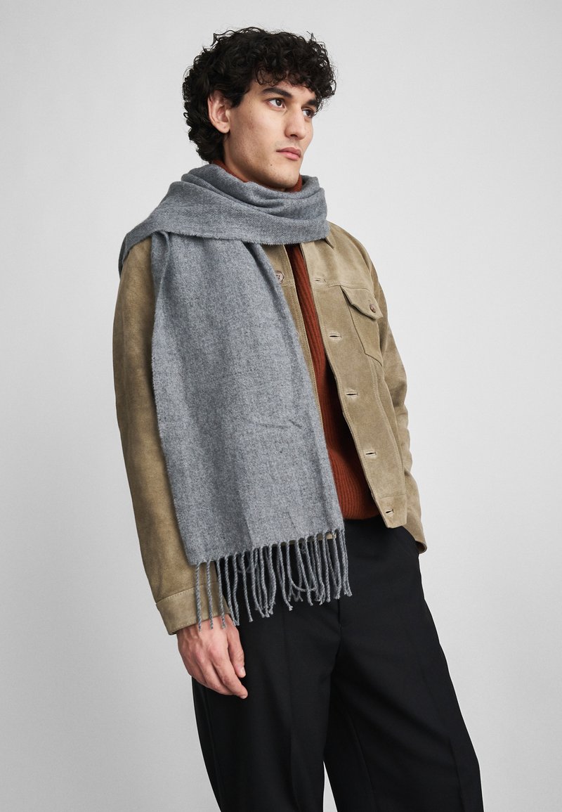 Γκρίζο μάλλινο κασκόλ με φραγκοειδείς άκρες, τοπ�οθετημένο γύρω από τον λαιμό, σε συνδυασμό με καφέ σουέτ μπουφάν και σκουρόχρωμο πουλόβερ.