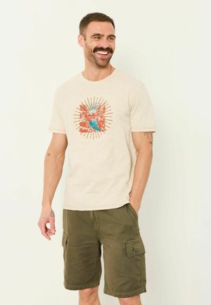 Lächelnder Mann mit Schnurrbart, der ein beiges Grafik-T-Shirt und olivgrüne Cargoshorts trägt, eine Hand in der Tasche, steht vor einem einfarbigen Hintergrund.