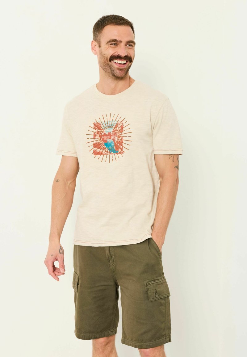 Homme souriant avec une moustache portant un t-shirt graphique beige et un short cargo vert olive, une main dans la poche, debout devant un fond uni.