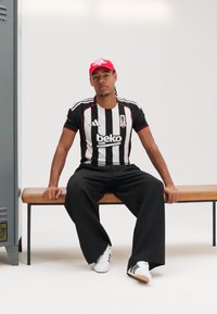 Must-valgete triipudega jalgpallivorm, millel on punased aktsendid, mustad laiad püksid ja valged tossud. Aksessuaaride hulka kuulub punane nokamüts.