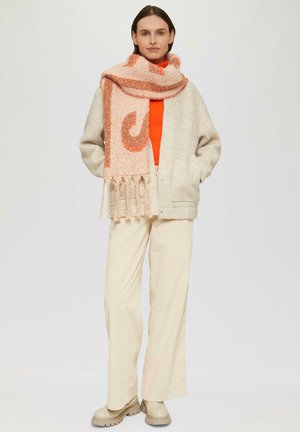 Lichtgrijze wollen jas, oranje coltrui, beige wijde broek en een roze patroon sjaal met franjes, staand tegen een neutrale achtergrond.