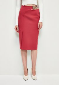 Jupe crayon rouge avec une fente à l'avant et une boucle carrée décorative à la taille, associée à des escarpins blancs à bout pointu. Texture de tissu lisse.