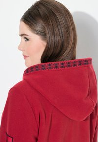 Punainen fleece-huppari, jossa on kuviolliset mustat yksityiskohdat hupussa. Pehmeä rakenne, joka tarjoaa rentoa istuvuutta ja pyöristetyn helman.