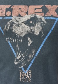 Donkergrijze T-shirt met een verweerd grafisch ontwerp van een T. rex-kop, met blauwe driehoekige accenten en tourdetails in vervaagd roze tekst.