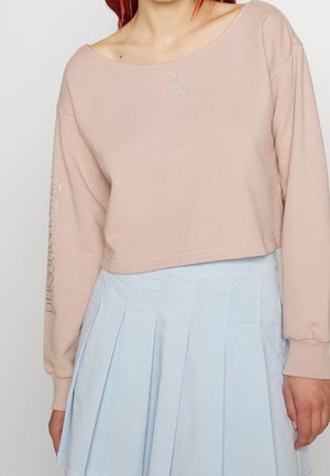Frau trägt ein hellrosa kurzes Sweatshirt mit Strass-Logo und einem hellblauen plissierten Rock vor einem schlichten Hintergrund.