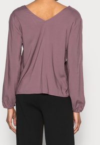Mujer vistiendo una blusa larga de manga larga en color malva con un escote en la espalda en forma de V y pantalones negros, vista desde atrás contra un fondo sencillo.