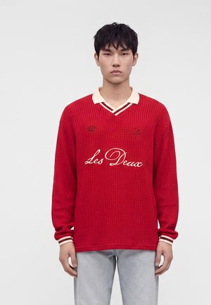 Young man with short black hair wearing a red textured sweater with "Les Deux" text, white collar, and light gray pants, standing against a plain background.