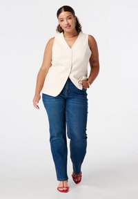 Beige mouwloze vest met knopen aan de voorkant en een getailleerde pasvorm, gecombineerd met blauwe denim jeans en rode sandalen met bandjes. Gladde stoftextuur.