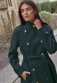 Zeleni dvoredni kaput s zlatnim dugmadima, visokim ovratnikom, pasom s remenom i bočnim džepovima, izrađen od mekog, teksturiranog materijala.
