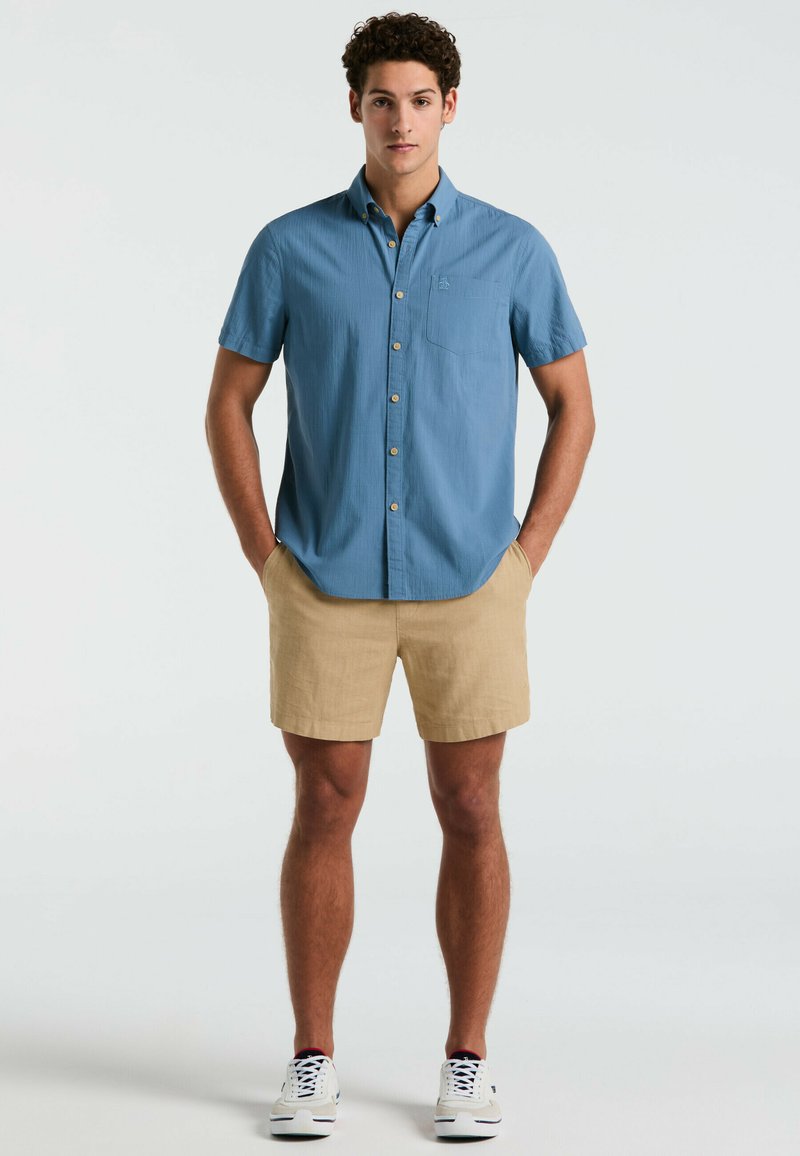 Korte mouwen, lichtblauwe overhemd met knoopsluiting en een borstzak, gecombineerd met beige shorts en witte sneakers. De stof heeft een gestructureerd patroon.