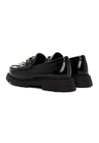 Mocasines de cuero negro con un acabado brillante, suela gruesa de goma negra, flecos decorativos y detalles en tono dorado.