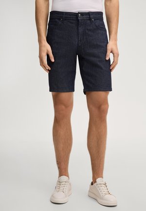 Mann trägt dunkelblaue Jeansshorts und weiße Turnschuhe, steht vor einem schlichten hellen Hintergrund.