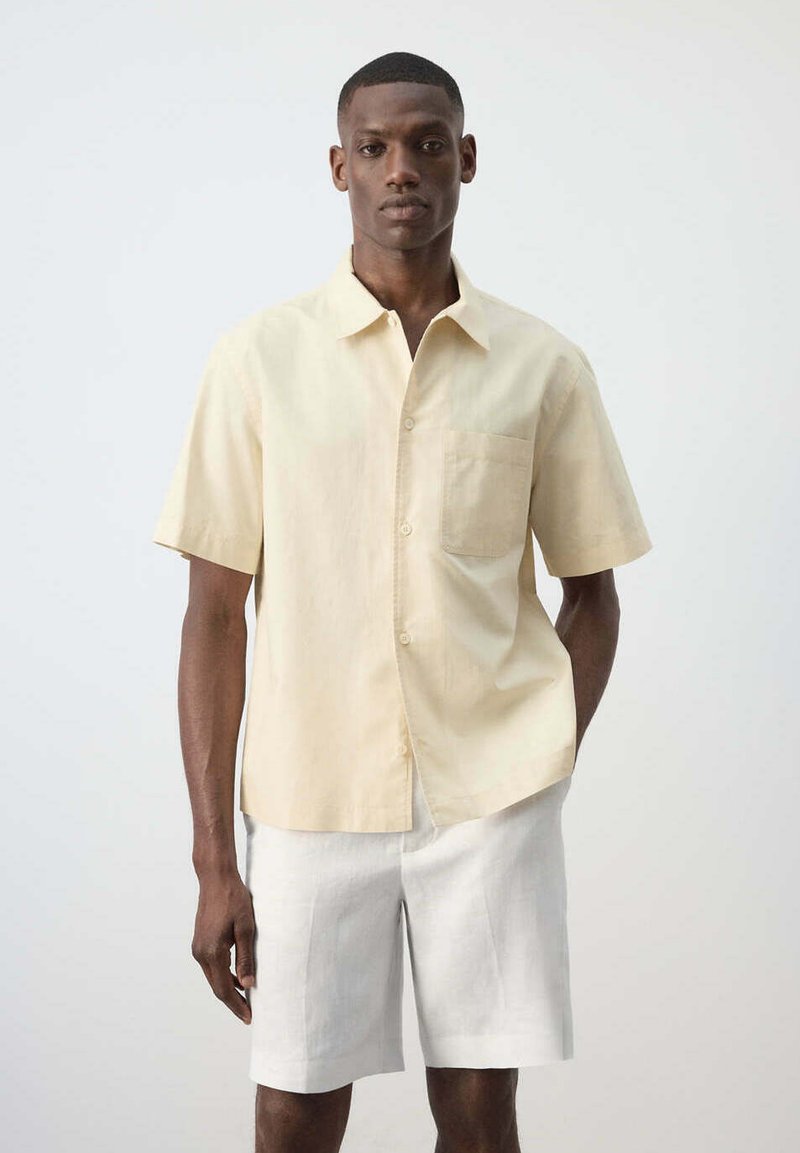 Homme portant une chemise beige à manches courtes avec poche poitrine et un short blanc aux genoux, debout devant un fond clair uni.