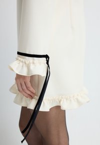 Abito color crema con maniche a volant e dettagli in velluto nero, rifinito con un nastro. Presenta un tessuto liscio e strutturato.