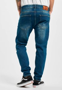 Jeans de denim oscuros de corte ajustado con un lavado deslavado, cinco bolsillos y un parche de cuero marrón en la cintura trasera. Combinados con zapatillas negras.