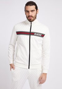 Hvid zip-up jakke med sorte detaljer, rød- og sorte striber samt "GUESS" logo. Fremstillet af struktureret stof med en sporty pasform.