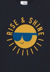 Granatowy sweter z motywem uśmiechniętego żółtego słońca w niebieskich okularach przeciwsłonecznych. Napis brzmi "RÓŚNĄ I BŁYSZCZ" w kolorze żółtym, otoczony promieniami słonecznymi.