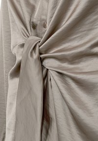 Beige satintyg som är samlat och knutet över en knappad skjorta, vilket framhäver texturen och de mjuka veckarna i materialet.
