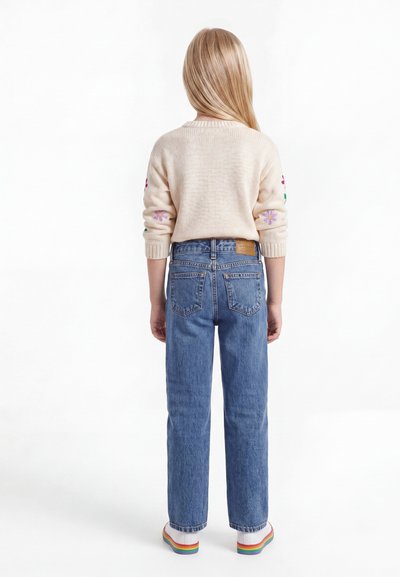 Παιδί με μακριά ξανθά μαλλιά που φοράει εκρού πουλόβερ με κεντήματα λουλουδιών, μπλε τζιν και λευκά παπούτσια με πολύχρωμες σόλες, στέκεται γυρισμένο από πίσω.