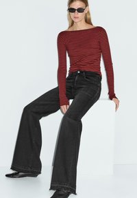 Maglia a maniche lunghe a righe rosse e nere, abbinata a jeans neri a vita alta a campana con orli grezzi. Modella seduta su un piedistallo bianco.