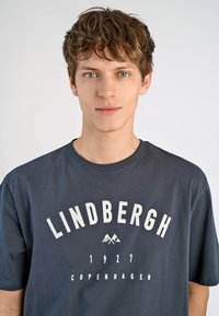 T-paita tummanharmaata puuvillaa, jossa on rohkeaa valkoista tekstiä "LINDBERGH 1927 KOPENHAGENA" ja vuoristokuvasto, pyöreä kaula-aukko ja lyhyet hihat.