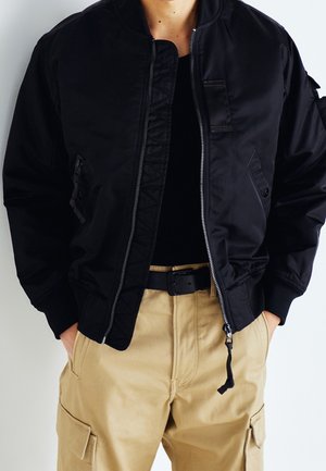 Person trägt eine schwarze Bomberjacke mit Reißverschluss über einem schwarzen Shirt, beige Cargohosen und einen schwarzen Gürtel, Hände in den Taschen.
